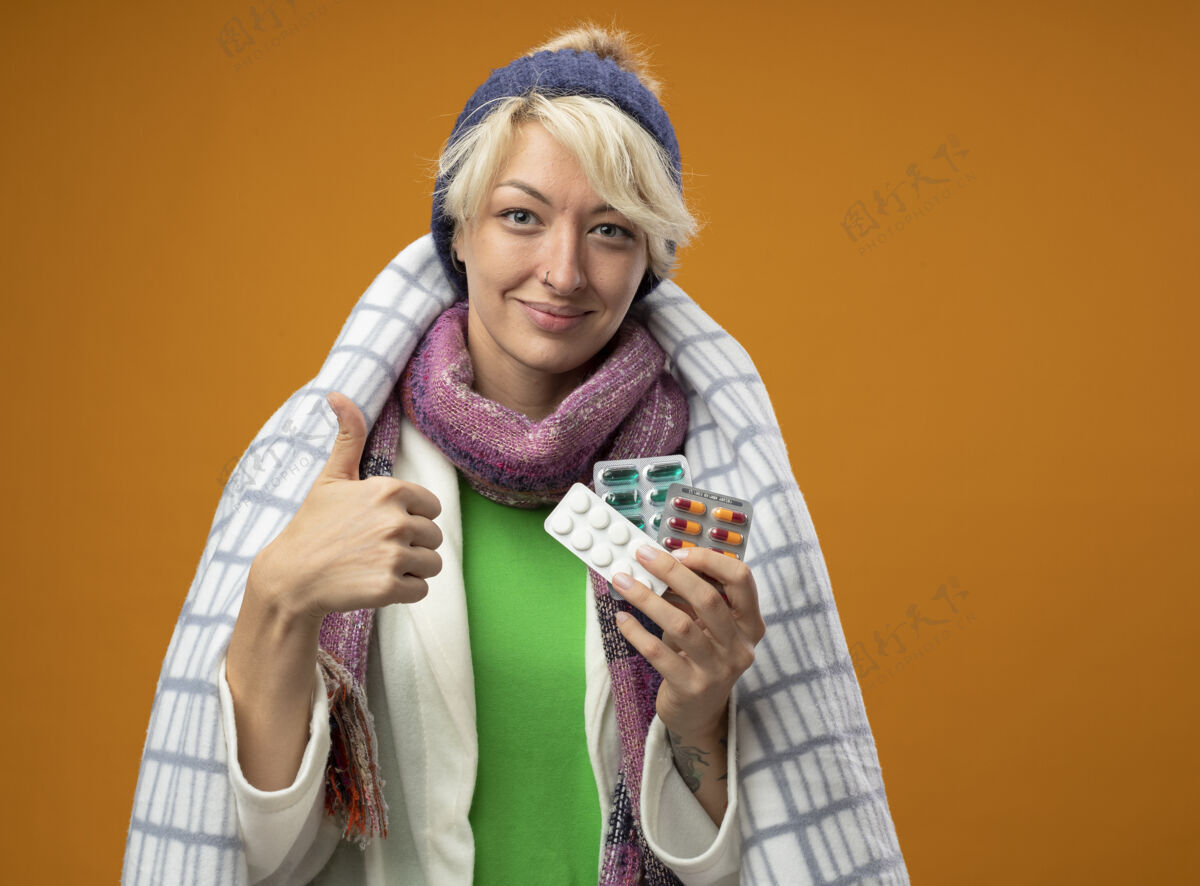 感觉生病的不健康的女人 短发披着暖和的围巾 帽子裹着毯子 手里拿着药片 微笑着竖起大拇指 站在橙色的墙上感觉好多了拇指温暖头发
