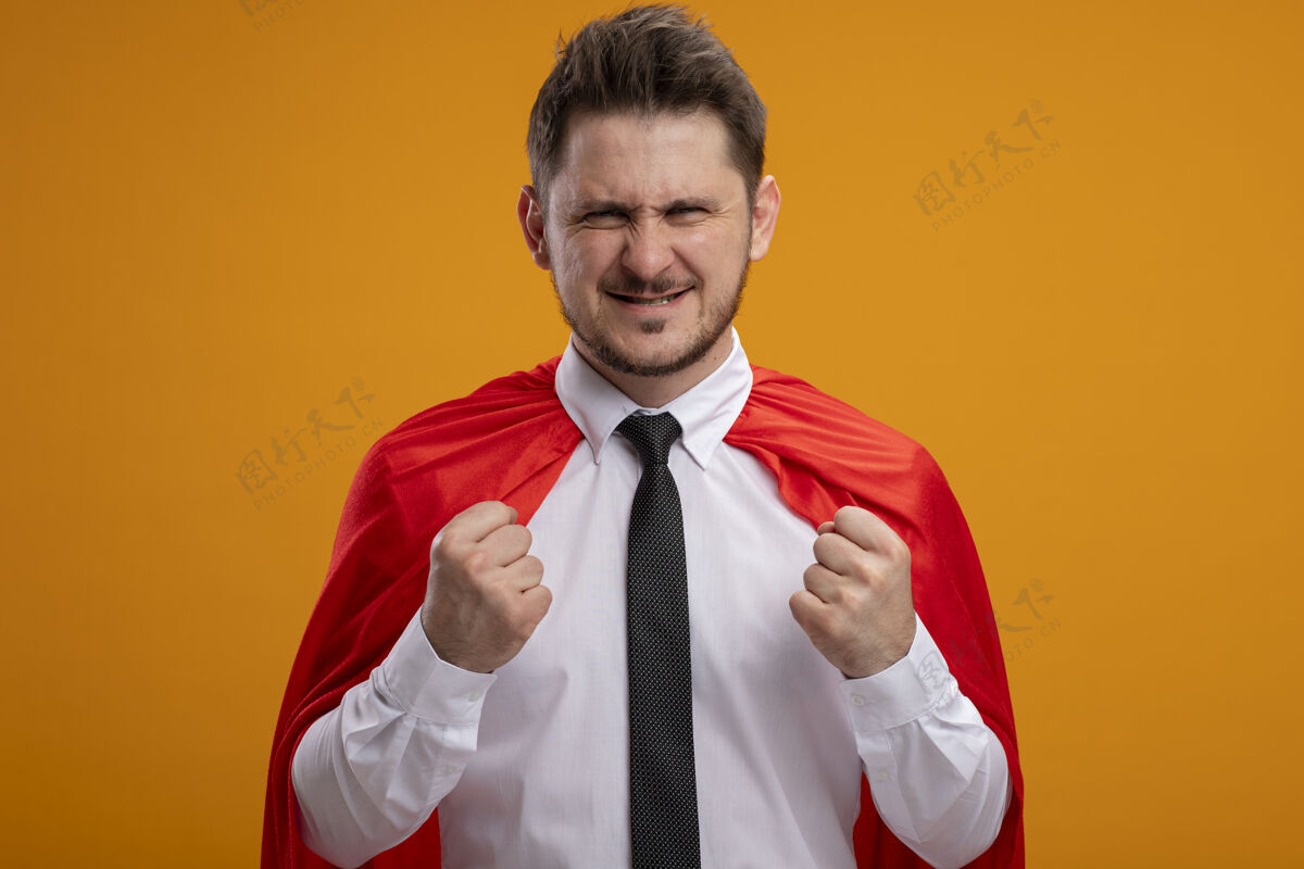 不高兴身穿红色斗篷的超级英雄商人站在橙色的墙上握紧拳头表示不满握紧超级立场
