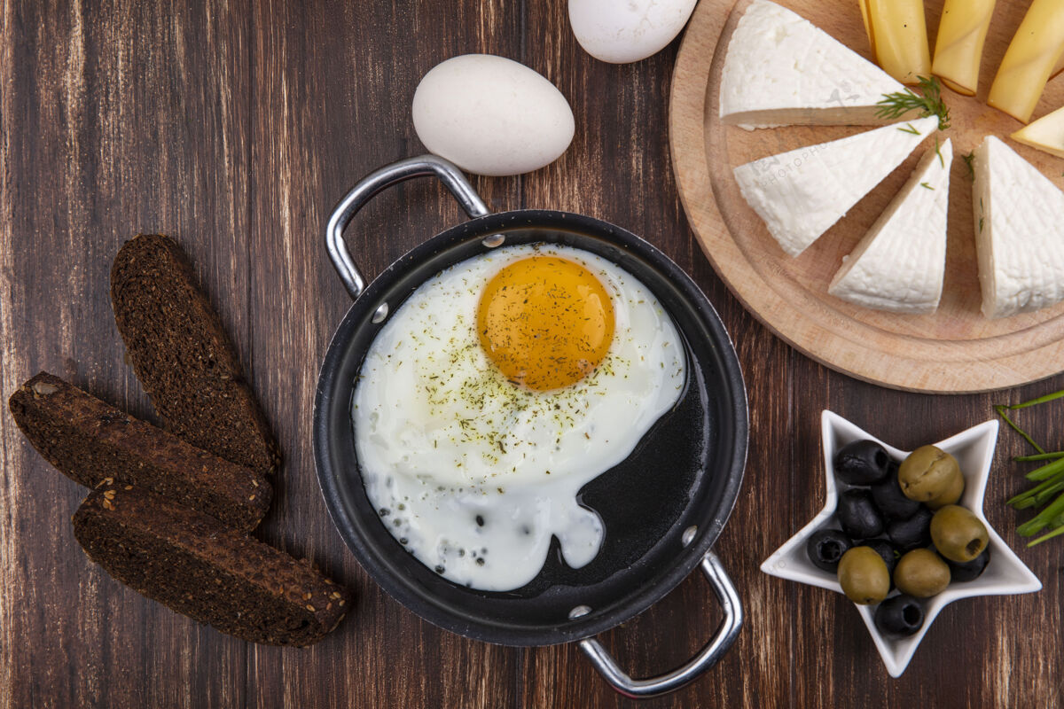 薯条俯视图：煎锅里的煎蛋 有橄榄 葱 黑面包和羊奶干酪 背景是木制的食物烹饪洋葱