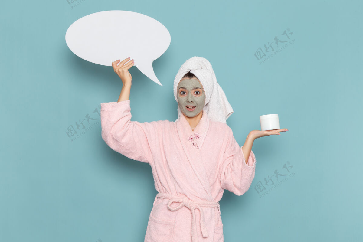 自理正面图：年轻女性 穿着粉色浴袍 淋浴后手持巨大的白色标牌 蓝色桌面上有美容水霜自助淋浴浴室桌子年轻人微笑