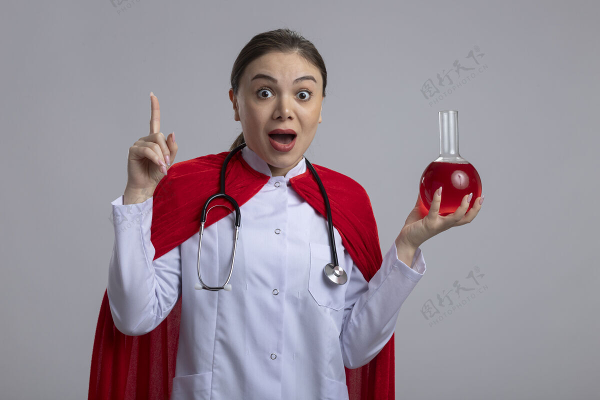 显示女医生手持听诊器 身穿白色医疗制服 身穿红色超级英雄斗篷 手持装有红色液体的烧瓶 显示食指有新想法 站在白色墙壁上兴奋而快乐手指医学举行