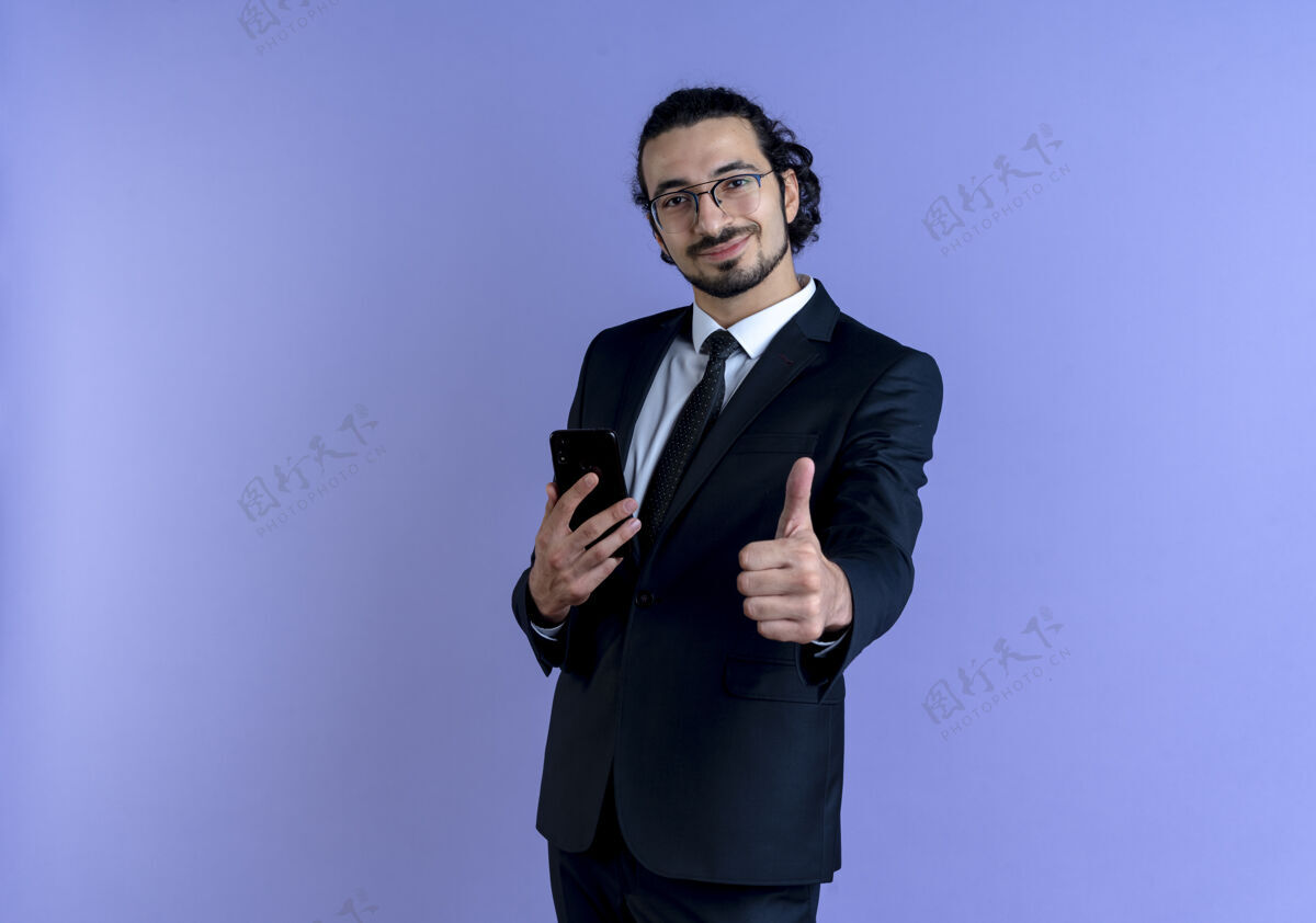 男士身穿黑色西装 戴眼镜 手持智能手机 竖起大拇指 自信地站在蓝色墙壁上的商人商务男士公司优雅