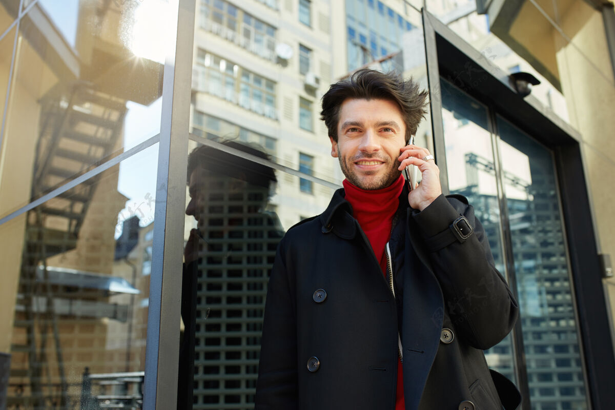 户外一张户外照片 年轻漂亮的黑发男人 穿着时髦的衣服 用智能手机打电话 面带愉快的微笑 积极地看着相机头发黑发毛衣
