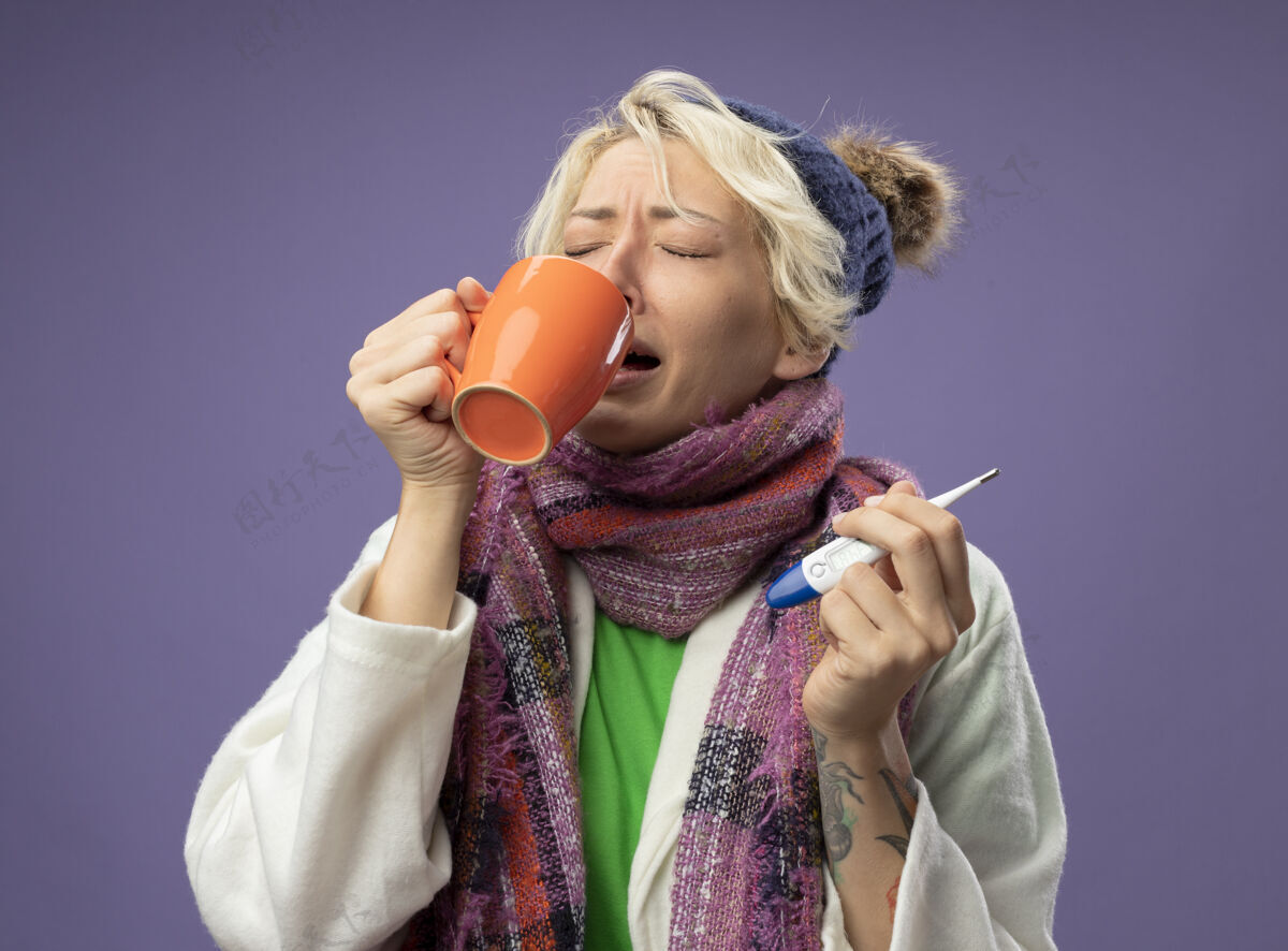 帽子生病的不健康的女人 短头发 戴着暖和的围巾和帽子 感觉不舒服 拿着一杯热茶和温度计站在紫色的墙上喝茶短裤体温计女人