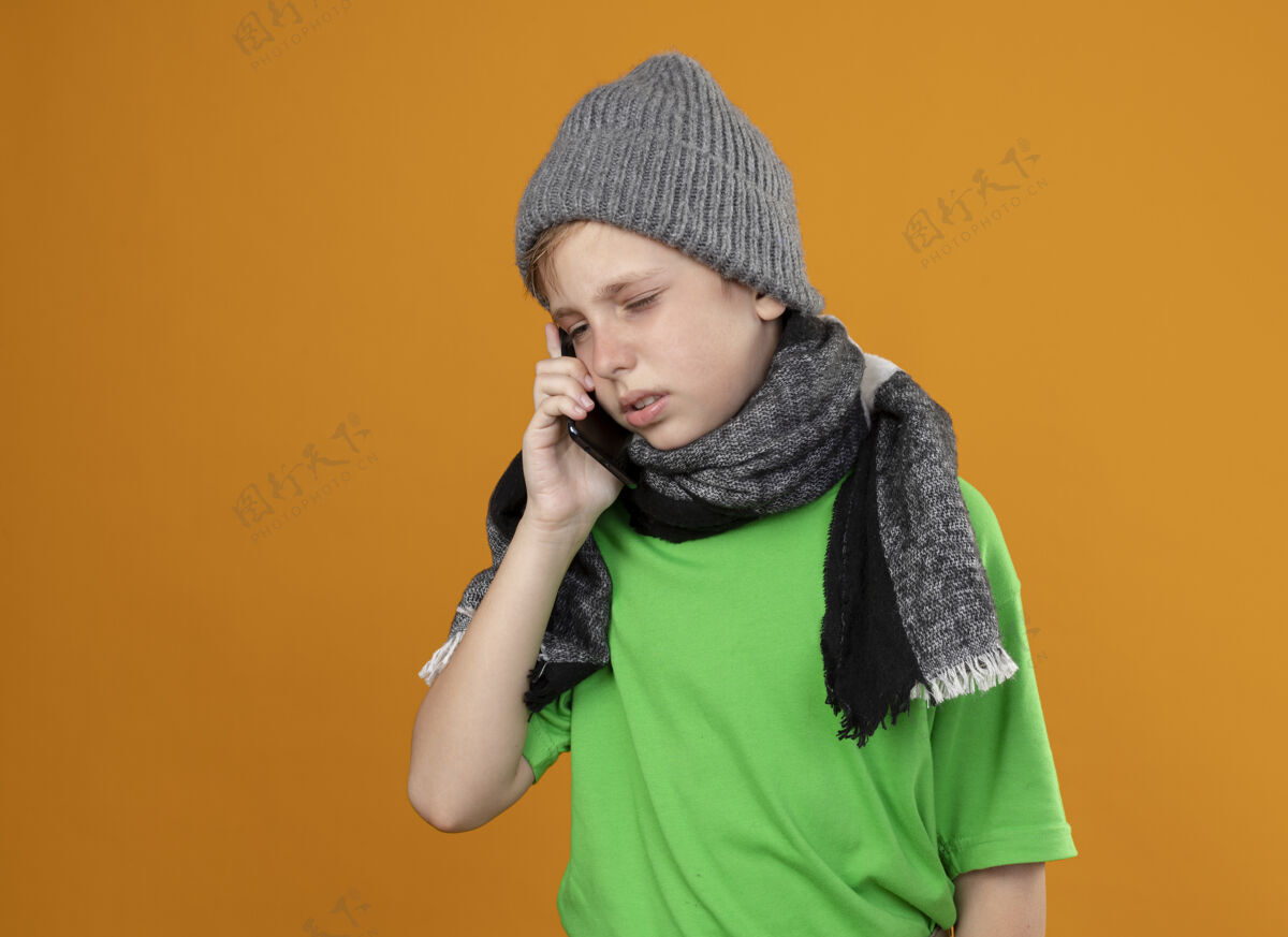 围巾生病的小男孩穿着绿色t恤 戴着暖和的围巾 戴着帽子 站在橘色的墙上打手机 感觉不舒服说话温暖帽子