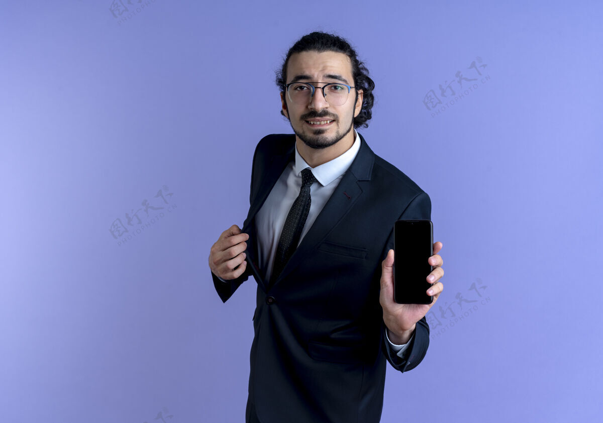 男人一个穿着黑色西装戴着眼镜的商人站在蓝色的墙上 向前方展示智能手机站优雅人