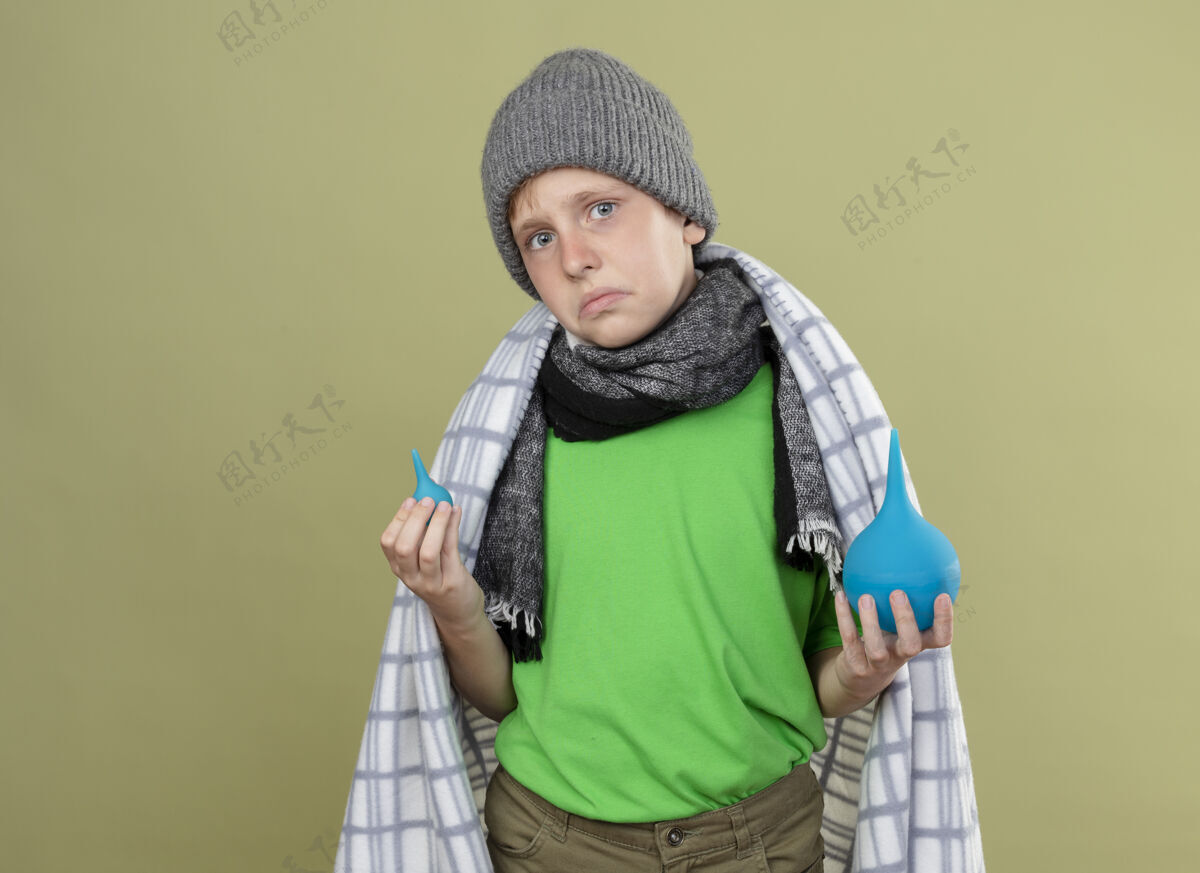 不高兴生病的小男孩戴着暖和的帽子 围着围巾 裹着毯子 拿着灌肠剂 站在轻薄的墙上 洛克感到困惑和不快温暖毯子少
