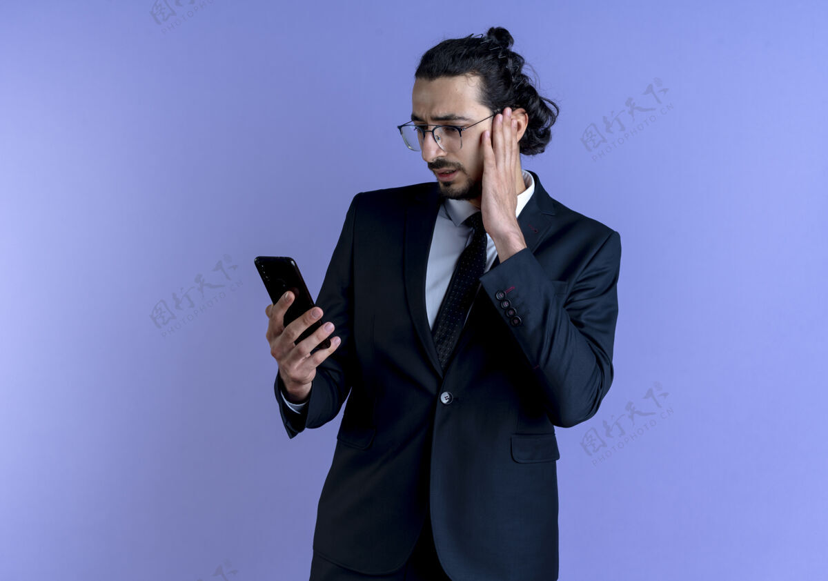 困惑一个穿着黑西装戴着眼镜的商人站在蓝色的墙上 用困惑的表情看着他的智能手机屏幕手机看商务