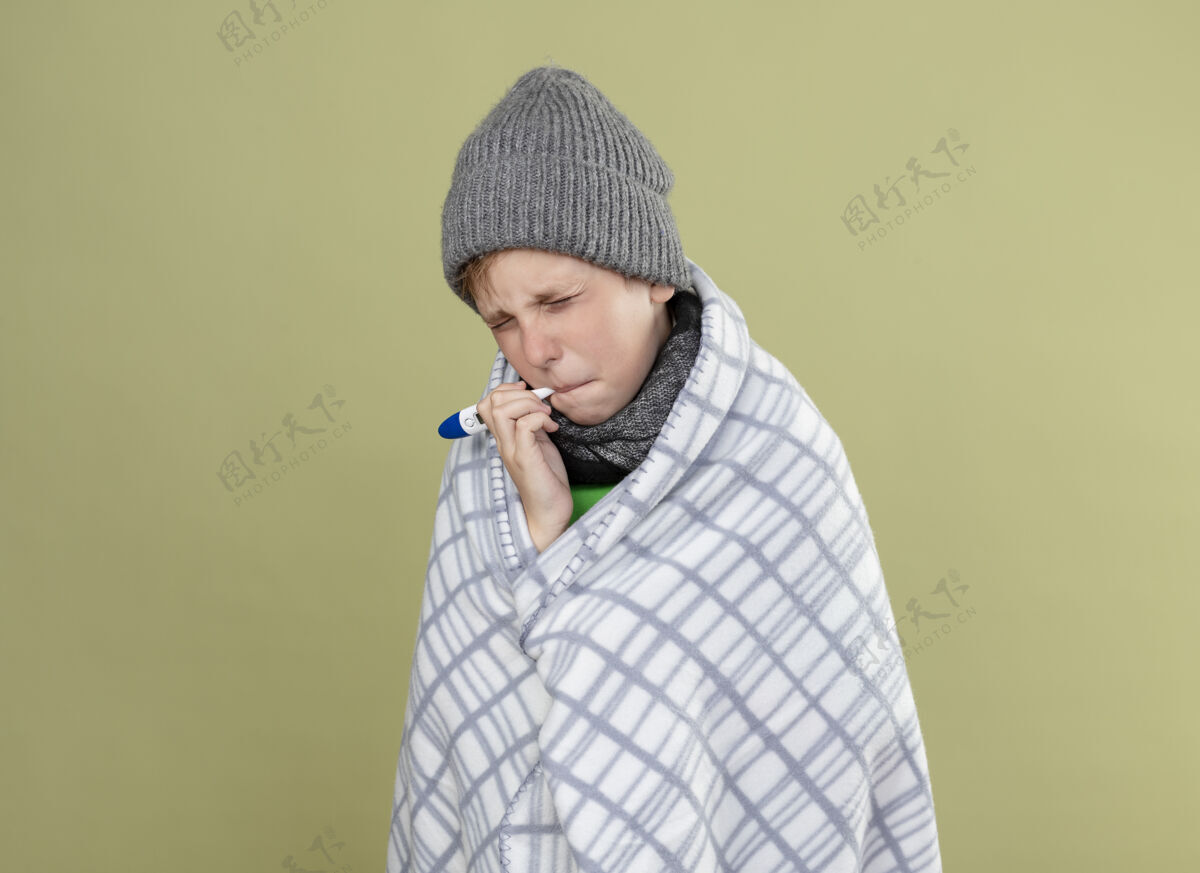 帽子生病的小男孩穿着绿色t恤 戴着暖和的围巾 帽子裹着毯子 把温度计放进嘴里 站在明亮的墙上生气地测量温度包男孩温度计