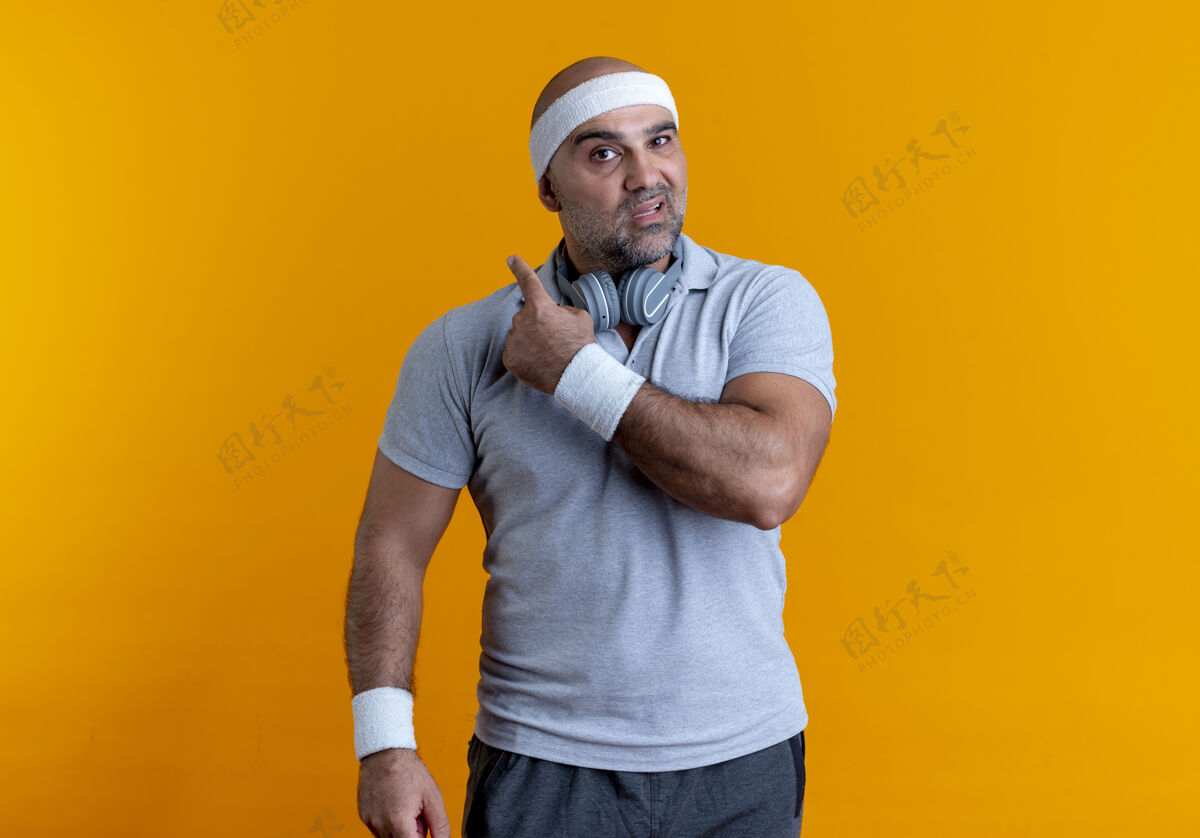 看戴着头巾的成熟的运动型男人站在橙色的墙上 用困惑的表情看着前面 指着后面运动教练运动员