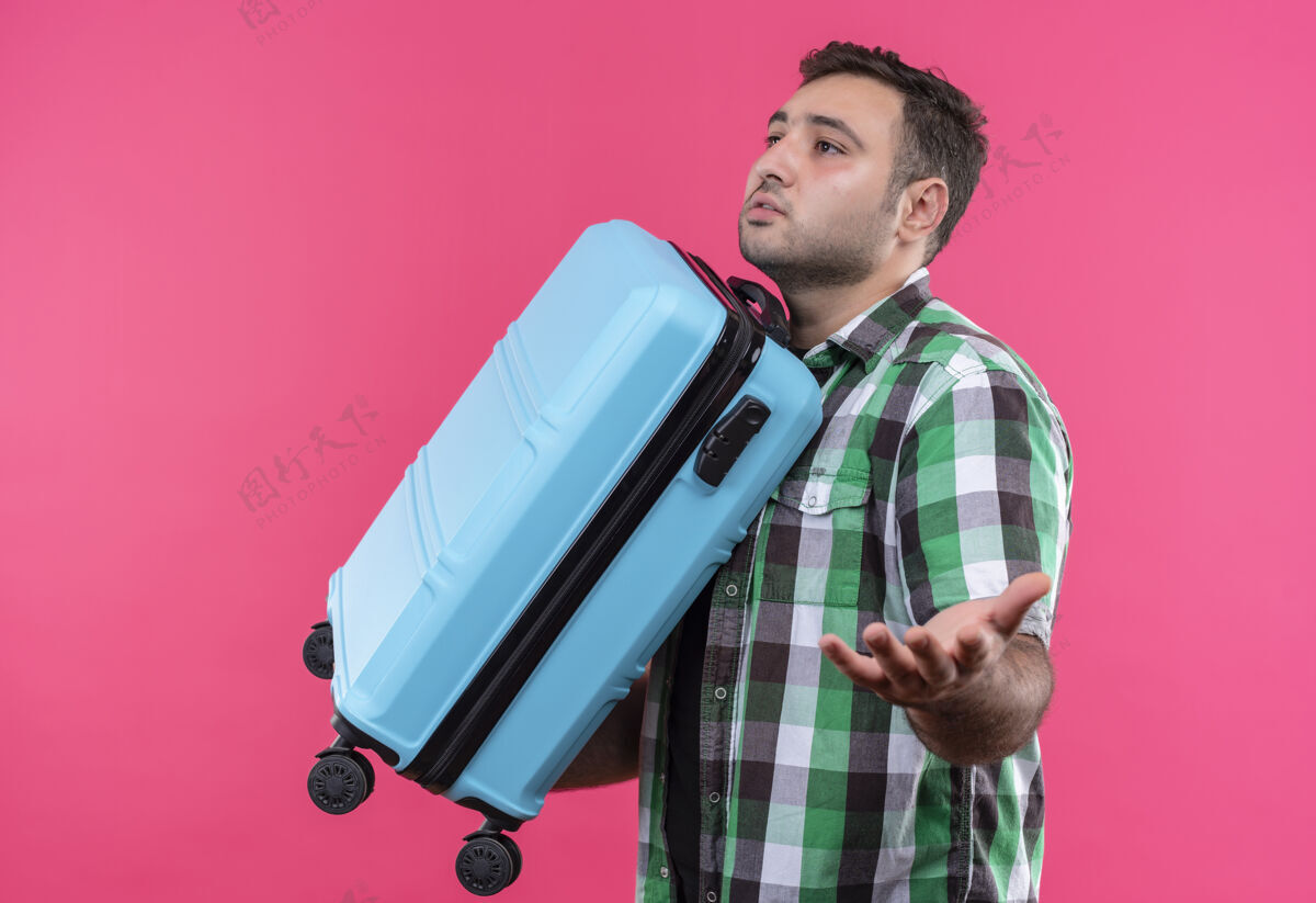 旅行穿着格子衬衫的年轻旅行家拿着手提箱 站在粉红色的墙上 茫然而不确定地看着一边男人看着困惑