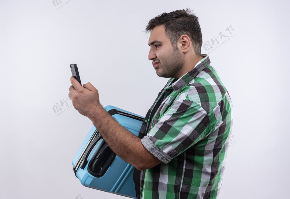 站穿着格子衬衫的年轻旅行家拿着手提箱站在那里看着智能手机的屏幕 满脸困惑地看着白色的墙壁姿势夏天假期