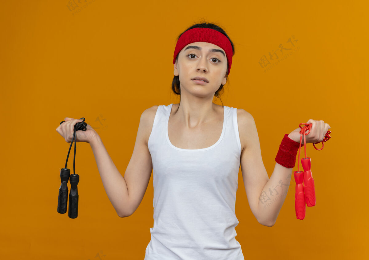 姿势身穿运动服的年轻健身女 头上系着两条跳绳 神情困惑 耸耸肩 站在橘色的墙上 心中充满疑惑运动员运动肩膀