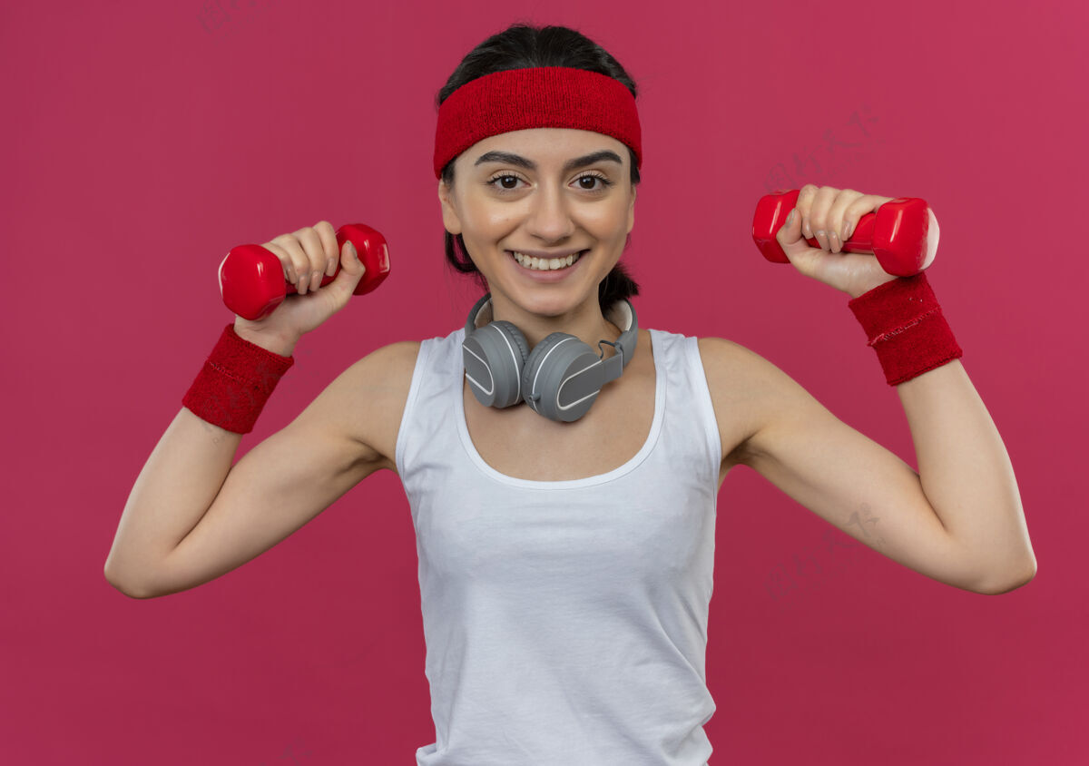 脸身穿运动服的年轻健身女 头戴钢带 举着两个哑铃 面带微笑地站在粉红色的墙上练习运动看人