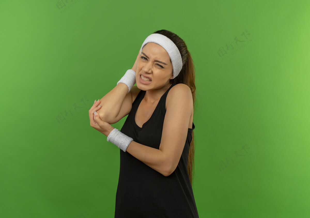 运动员穿着运动服的年轻健身女 头箍碰到手肘 站在绿墙上疼痛难忍年轻人人疼痛