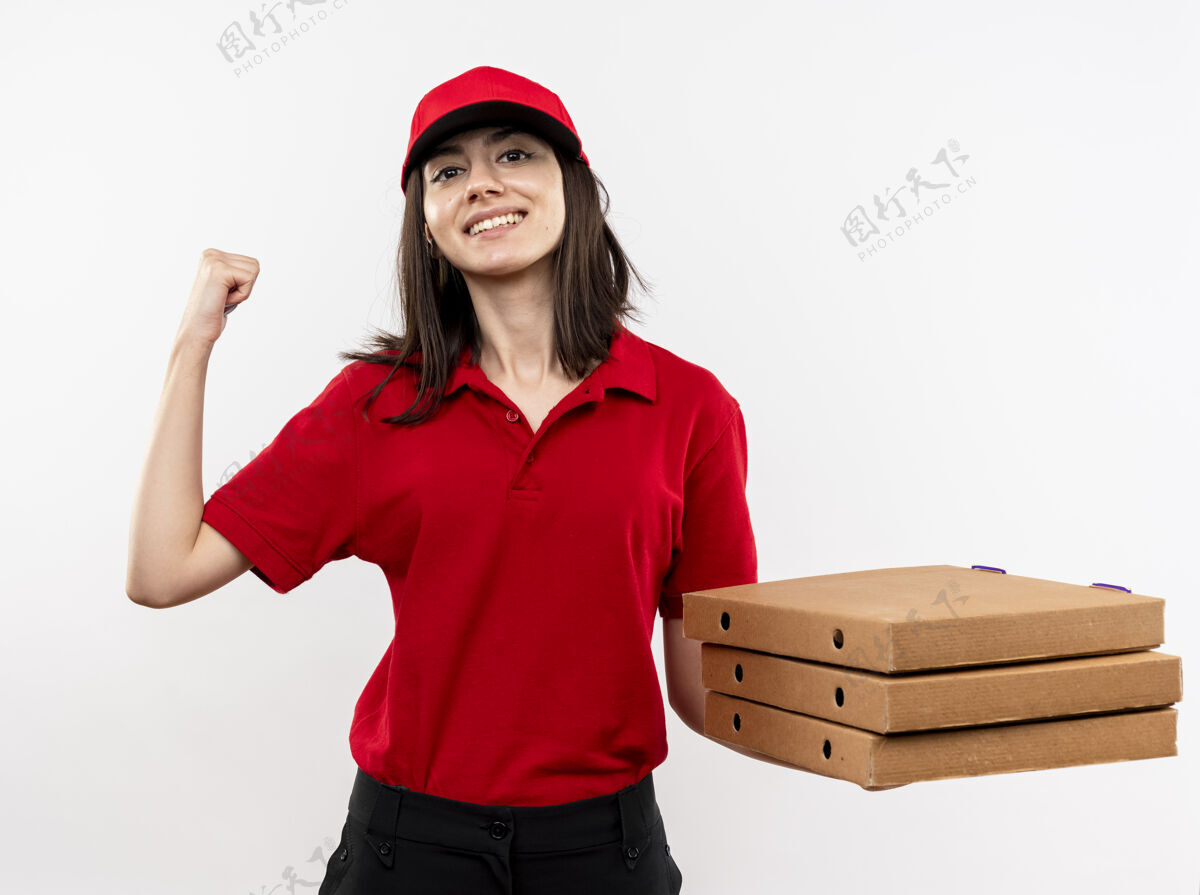 正面年轻的送货员身穿红色制服 头戴鸭舌帽 手里拿着一叠披萨盒 握紧拳头 站在白色背景上 开心而积极地微笑着年轻站着拳头