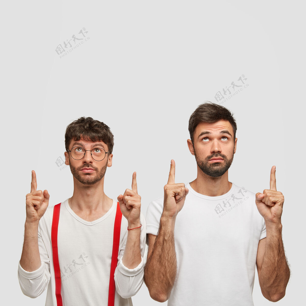 自由空间两个留着胡须的年轻人的照片 表情严肃 食指朝上 展示酷的复制空间 在白墙上做广告 穿着休闲服装促销未刮胡子手势