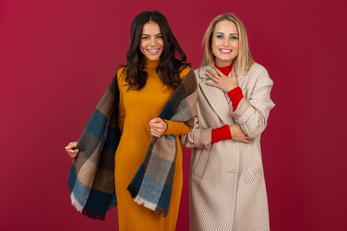 时尚两位笑容可掬的时尚女性穿着秋冬时装和外套 在红墙上孤零零地摆着姿势冷颜色毛衣