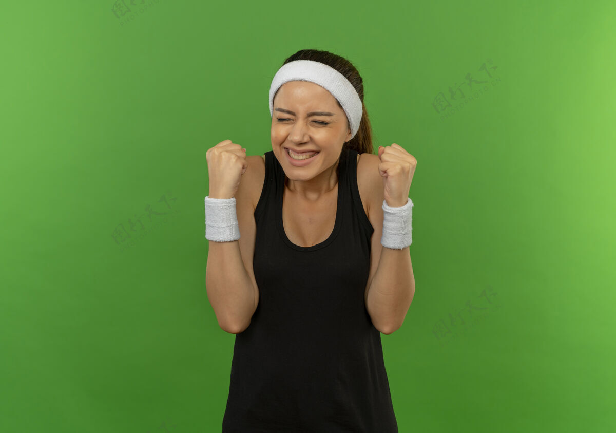 年轻身穿运动服 头箍紧握拳头的年轻健身女士站在绿色的墙壁上 既高兴又兴奋握紧运动装人