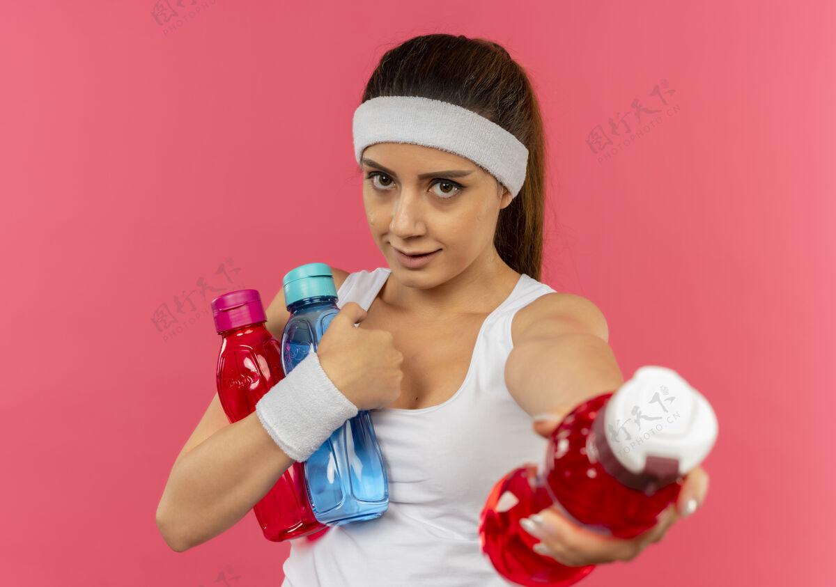 头带穿着运动服的年轻健身女 头上戴着一条带子 手里拿着几瓶水 站在粉红色的墙上 给其中一瓶水喝公民运动装持有