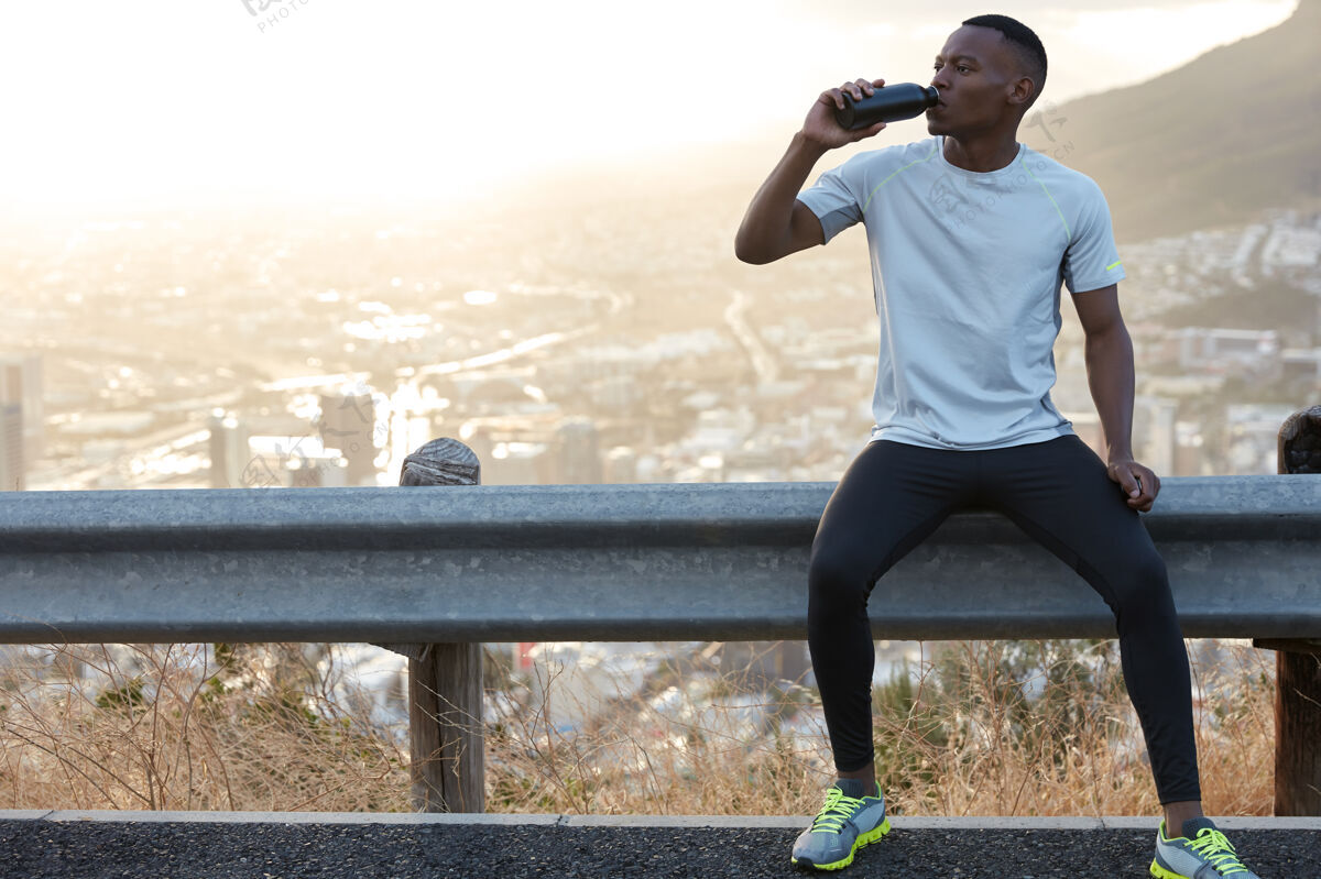 惊人非洲裔美国人口渴的人喝淡水 享受户外运动训练后的休息 坐在路标旁 风景优美的全景山景复制空间 用于宣传内容或信息活跃水合肌肉