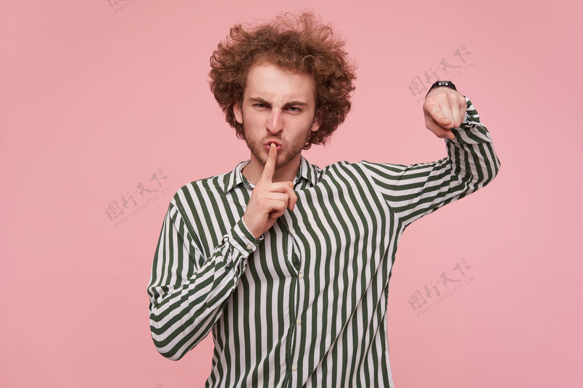 食指一张室内照片 穿着休闲衬衫的年轻卷发红发男子皱着眉头 把食指举在嘴唇上 靠着粉色的墙壁站着20多岁男人狐狸精
