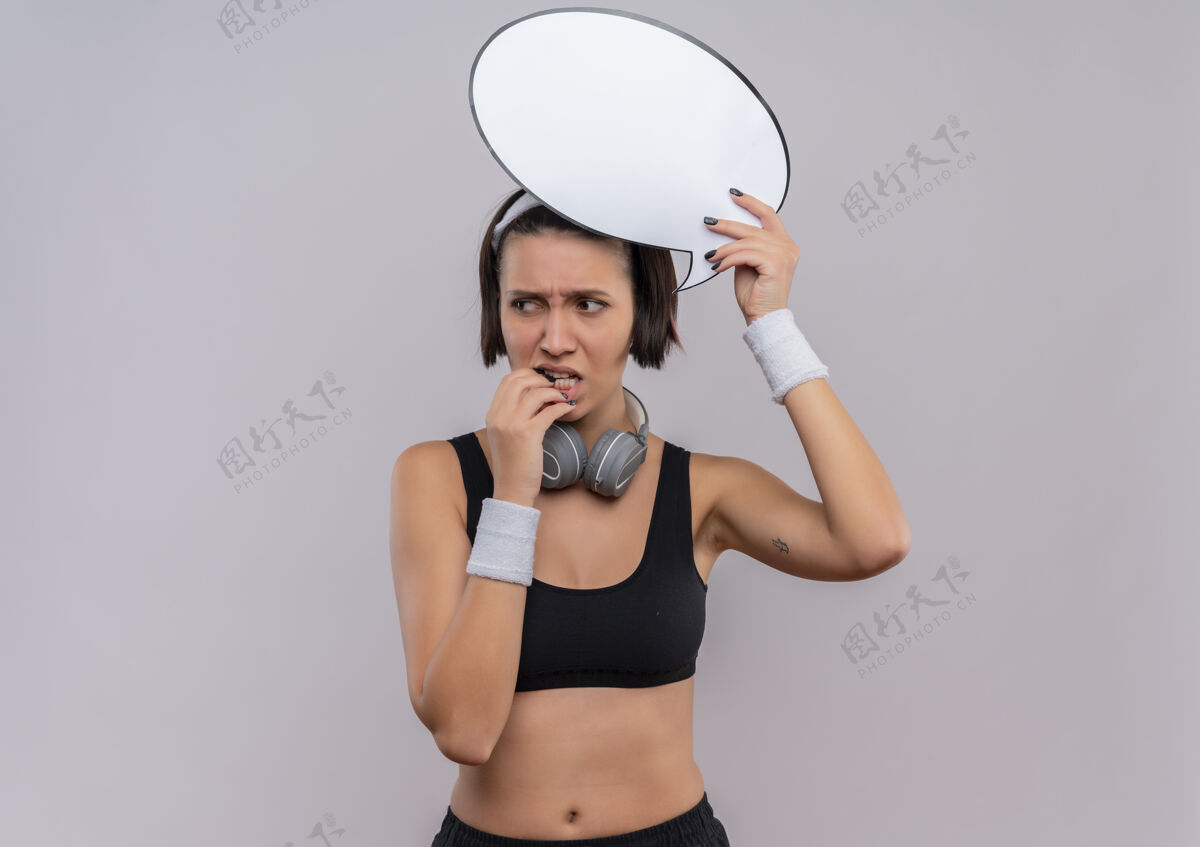 健身穿着运动服的年轻健身女 头上戴着一个空白的演讲泡泡牌 站在白色的墙上困惑地看着一边健康抱着运动