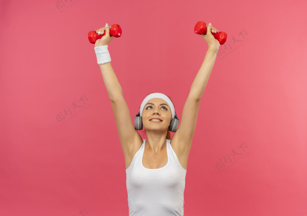 抱着身穿运动服 头箍 举着两个哑铃 站在粉色墙壁上做运动的年轻健身女士 看上去很自信人看运动
