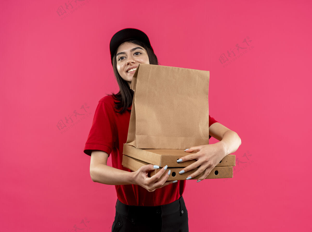 包装穿着红色马球衫和帽子的年轻送货女孩拿着纸包和披萨poxes站在粉色背景下 面带微笑地看着一旁微笑持有送货