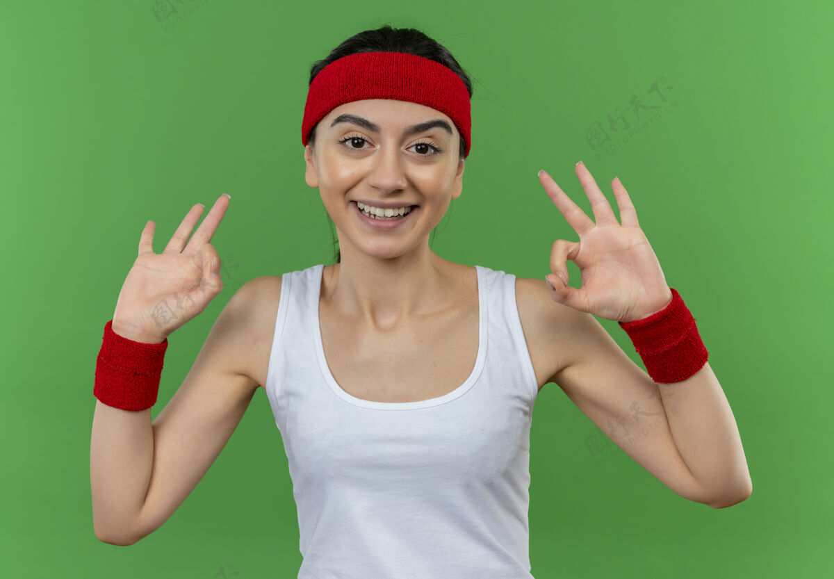 运动身穿运动服的年轻健身女士 头上戴着一条快乐的头巾 面带乐观的笑容 站在绿色的墙上 高高兴兴地做着“ok”的手势年轻人健身