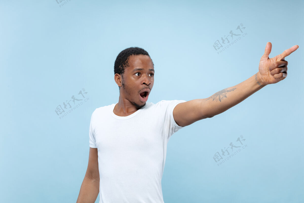 人在蓝色背景上 一个穿着白衬衫的非洲裔美国年轻人的半身特写肖像人类情感 面部表情 广告 销售理念指向 选择 惊讶复制空间年轻衬衫手势