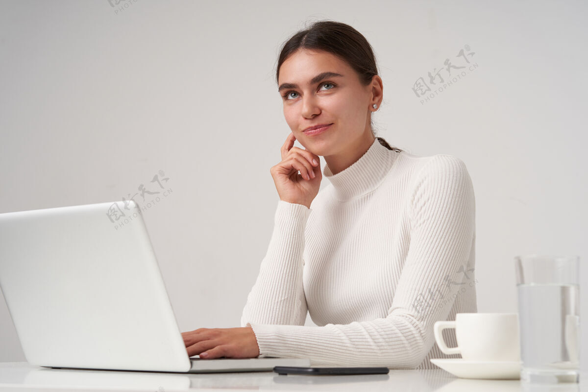思想室内拍摄的迷人的积极的年轻黑发女子一直举起手放在脸颊上 带着愉快的微笑梦幻般地看着一边 隔着白墙欧式笔记本电脑穿着
