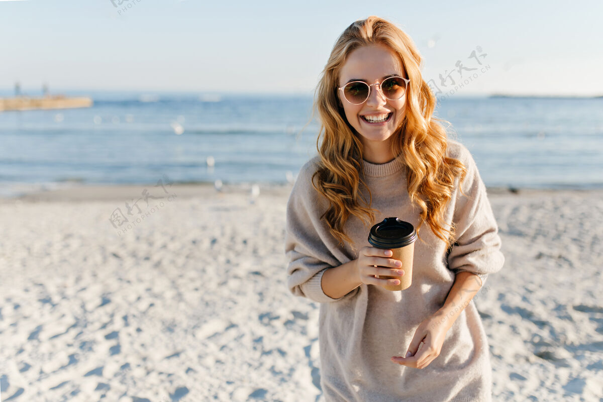 装备迷人的女人 卷发 在沙滩上喝茶时髦的女人 穿着毛衣 在沙滩上放松秋日天空假期金发