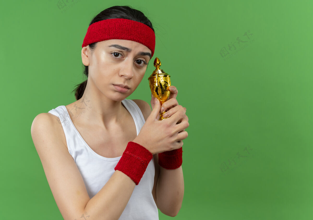 人身穿运动服 头戴钢带 手持奖杯 脸上带着悲伤表情的年轻健身女士站在绿色的墙上运动运动员站着
