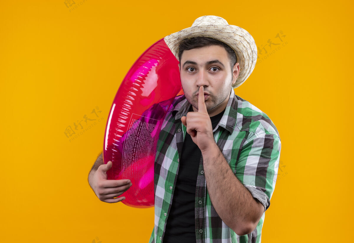 男人穿着格子衬衫 戴着夏帽 手持充气戒指 手指放在嘴唇上 站在橙色的墙上做着沉默的手势假期年轻人人