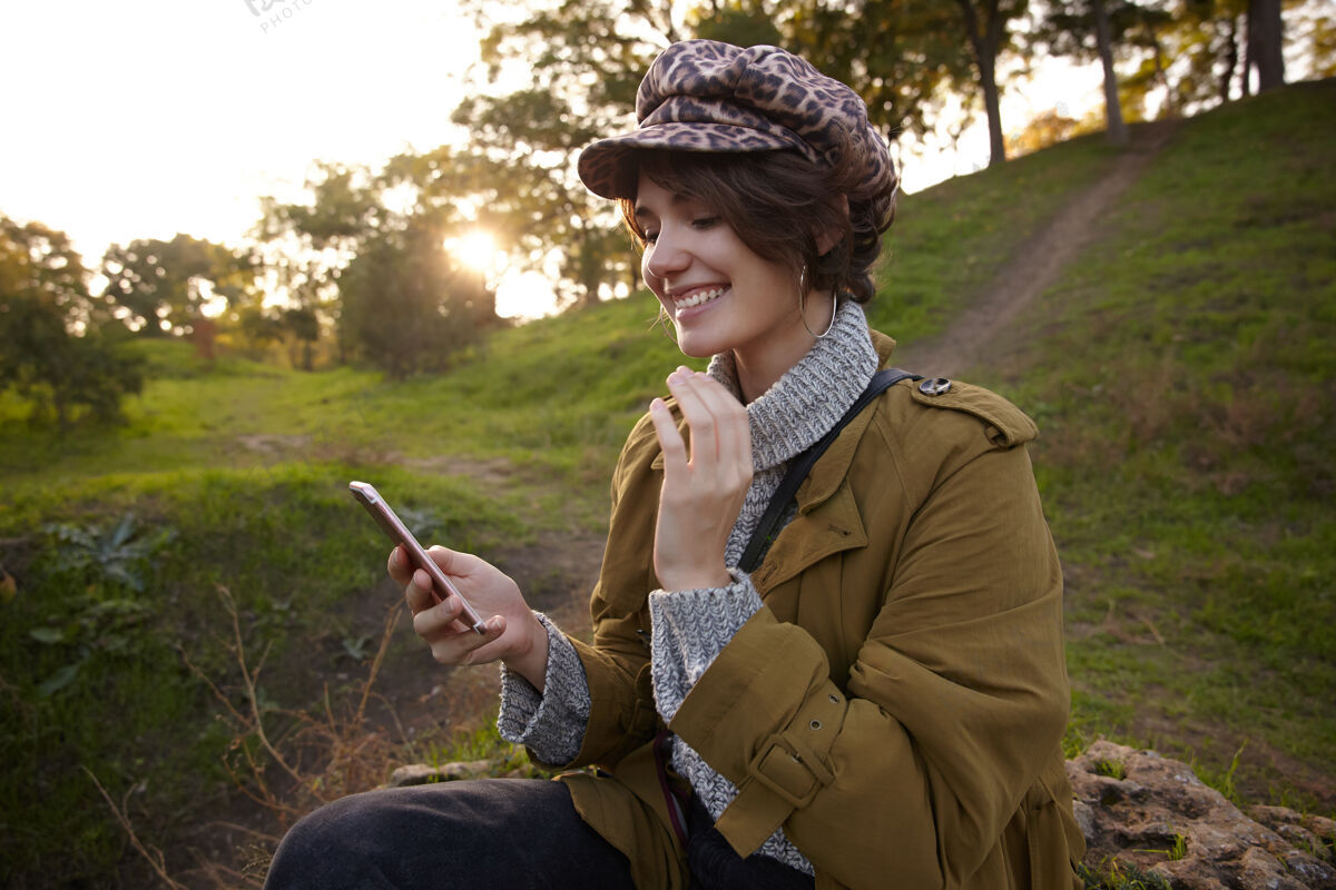 公园快乐的年轻漂亮的棕色头发的女性 鲍勃的发型 举起手拿着手机 读着朋友的留言 在夕阳下模糊的公园里摆姿势模特秋天女性