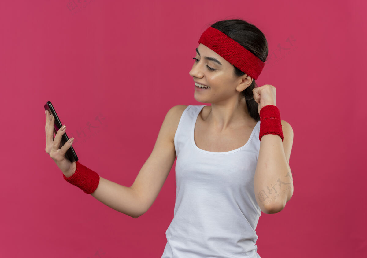 运动装身穿运动服的年轻健身女士 头箍紧挨着智能手机屏幕 微笑着握紧拳头站在粉色的墙上姿势微笑年轻人