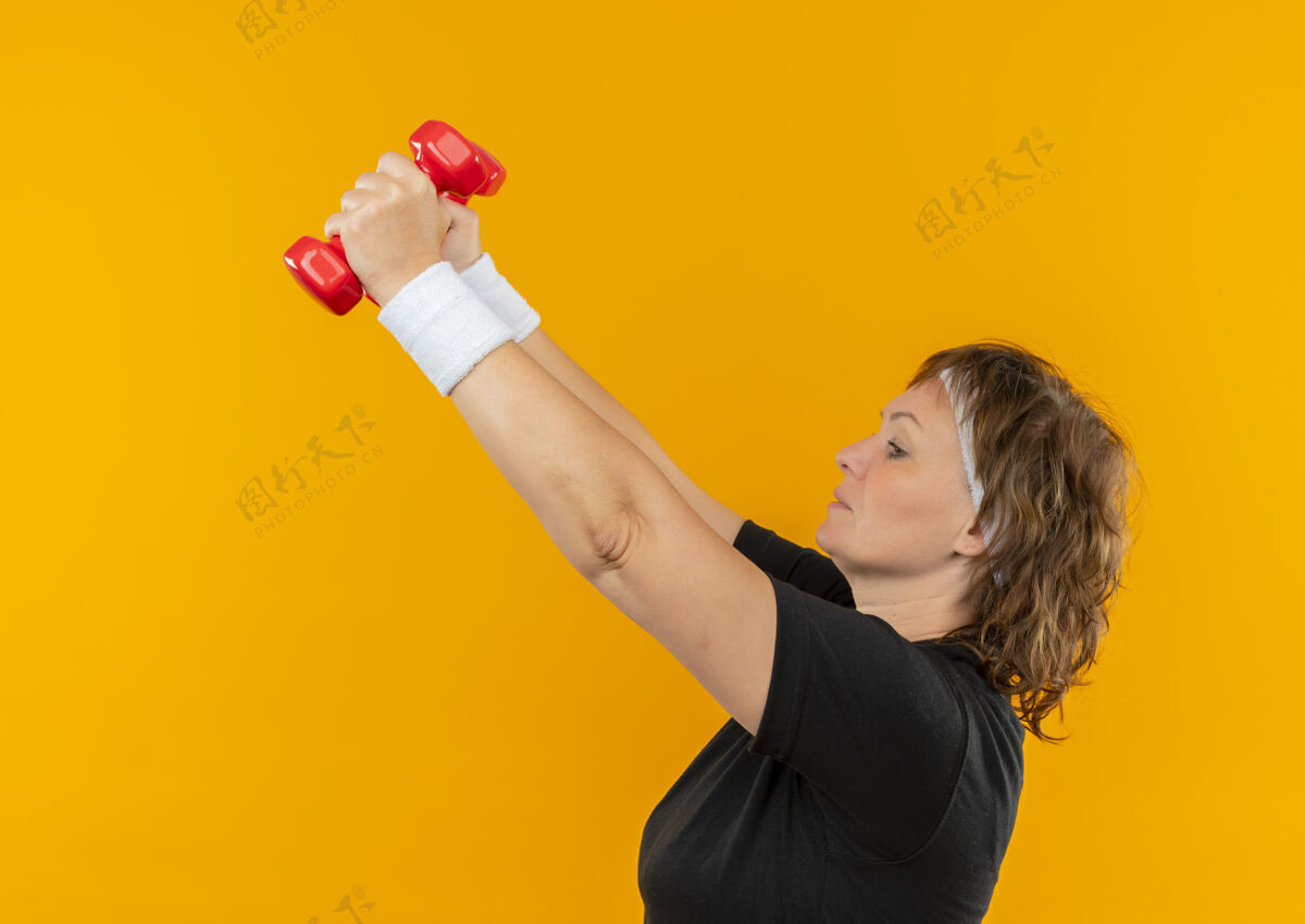 抬高中年运动女性 身穿黑色t恤 戴着头带 举着哑铃 站在橙色的墙上 看起来自信而紧张运动头带女人