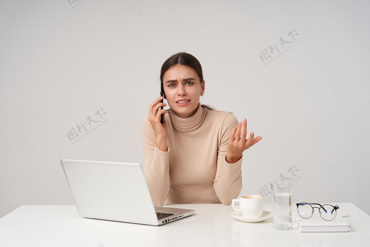 欧式困惑的年轻蓝眼睛黑头发的女士穿着正式的衣服 坐在办公室里用现代化的笔记本电脑 眉头紧锁 一边用不愉快的电话交谈头发优雅工作