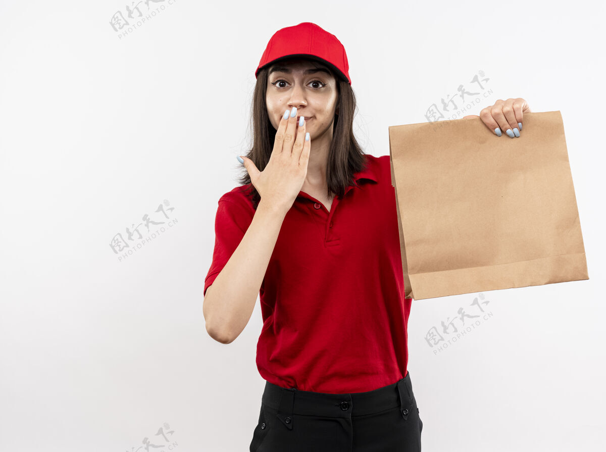 帽子年轻的送货女孩穿着红色制服 戴着帽子 拿着纸包看着镜头 惊讶地用手站在白色的背景上举行年轻立场