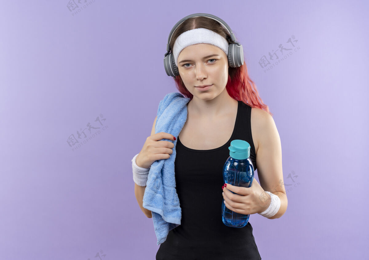 健身穿着运动服的年轻健身女孩戴着耳机 脖子上挂着毛巾 手里拿着一瓶水站在紫色的墙上人女孩毛巾