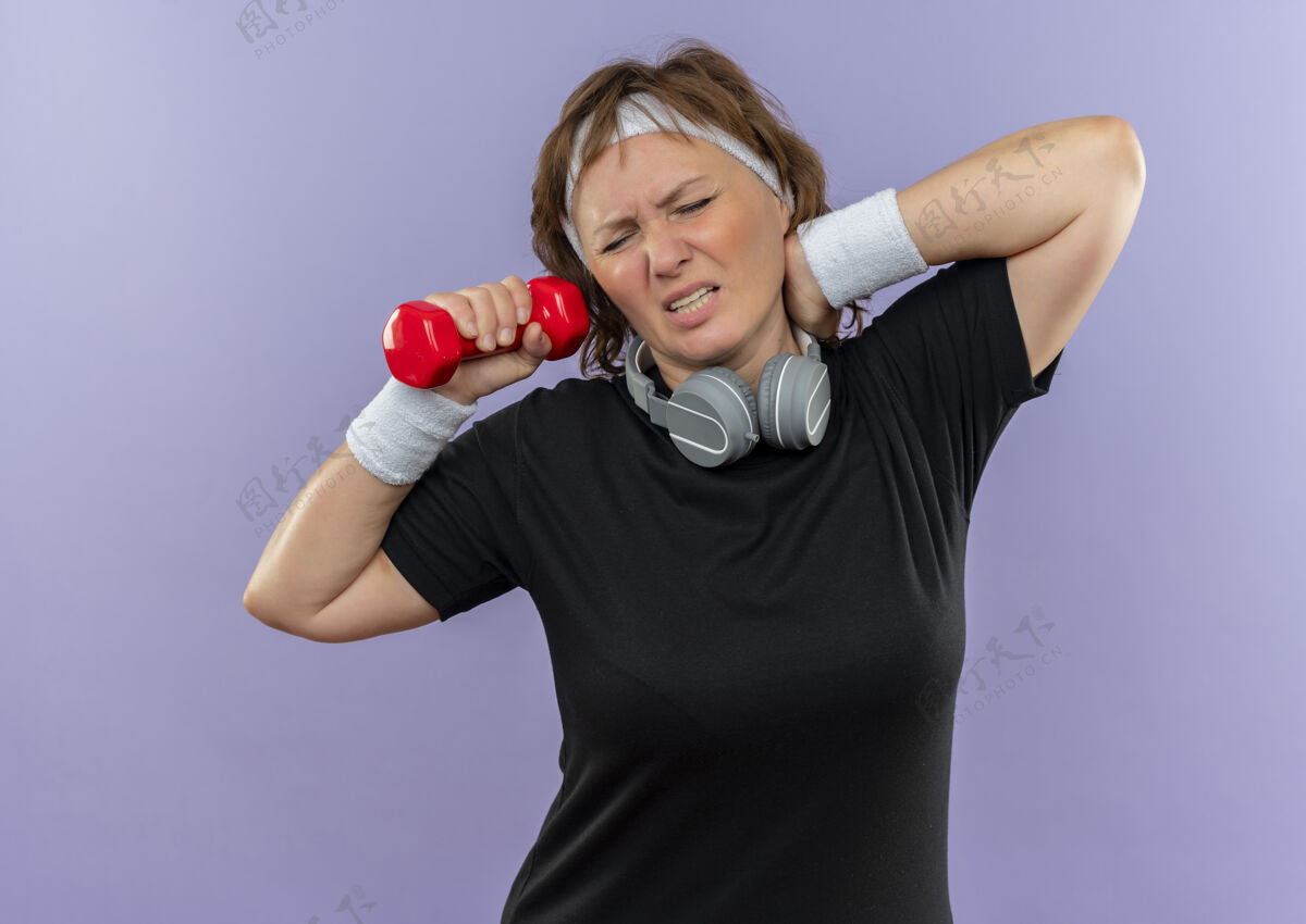 触摸中年运动女性 身穿黑色t恤 戴着头带 戴着哑铃 看起来很累 筋疲力尽 摸着脖子 站在蓝色的墙上感觉疼痛运动员疲惫头带