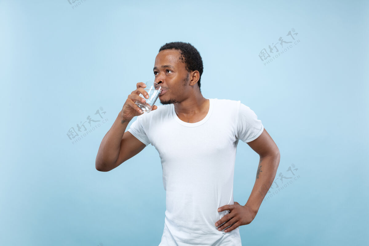 面部在蓝色背景上 一个身穿白衬衫的非洲裔美国年轻人的半身特写肖像人类的情感 面部表情 广告概念拿着一个玻璃杯和饮用水行政人员人公司
