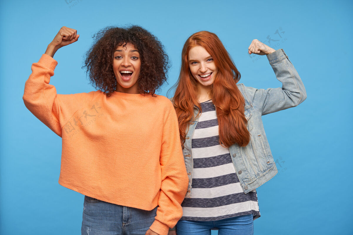 快乐快乐迷人的女士们开心地看着 开心地笑着 举起双手 同时展示强壮的肱二头肌 隔离在蓝色的墙壁上卷发毛衣女性