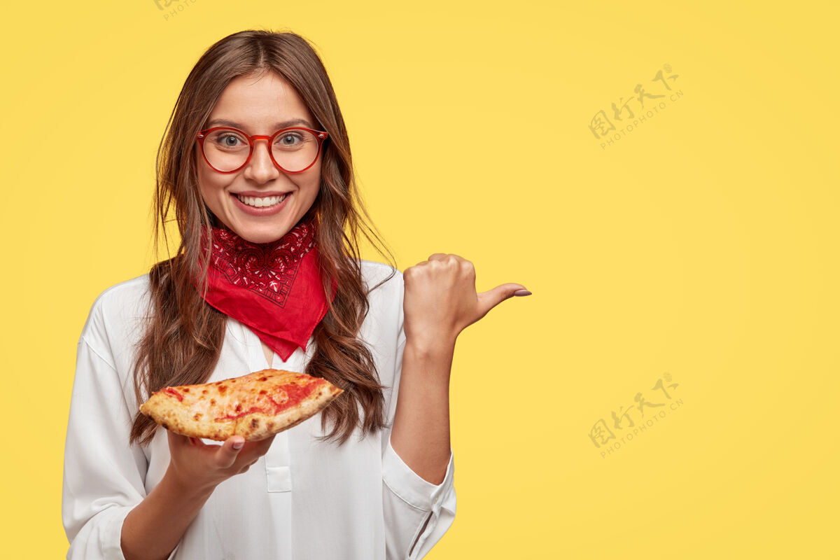 午餐高兴的微笑着的女人拿着美味的比萨饼 用拇指指了指她买比萨饼的地方 在比萨店做广告 戴着红色的头巾和白色的衬衫 隔着黄色的墙高兴积极凝视