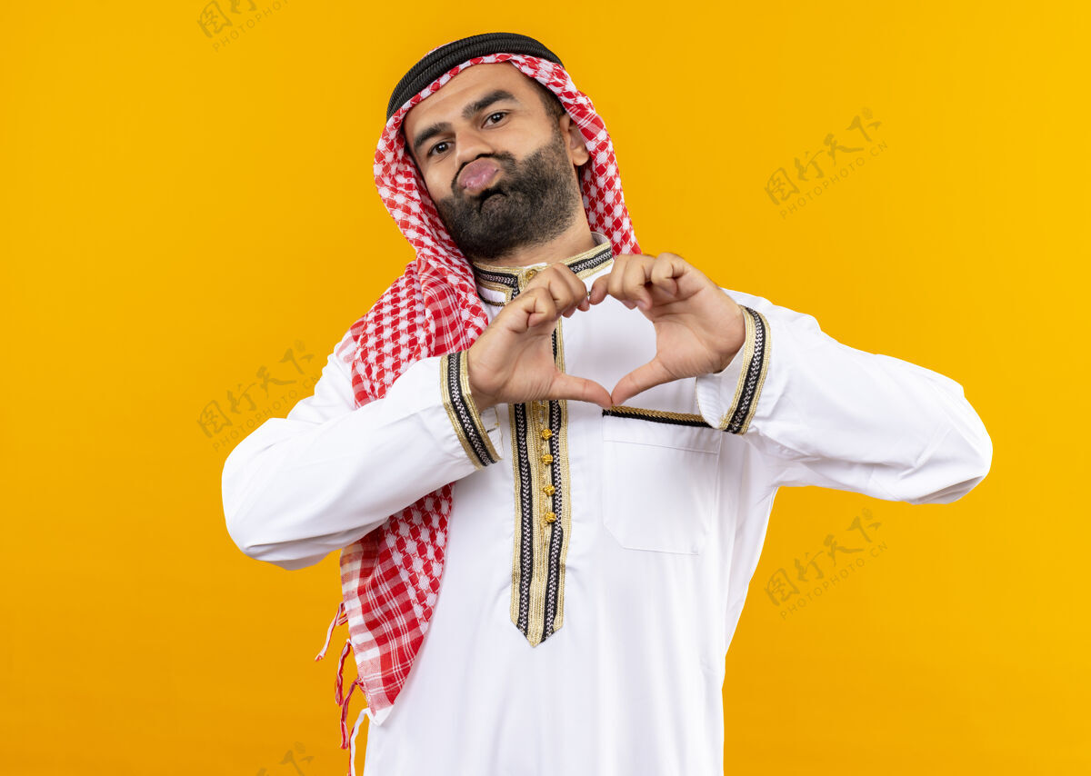 站身着传统服装的阿拉伯商人站在橙色的墙上 用手指在胸前做着一个亲吻的手势使胸部尝试