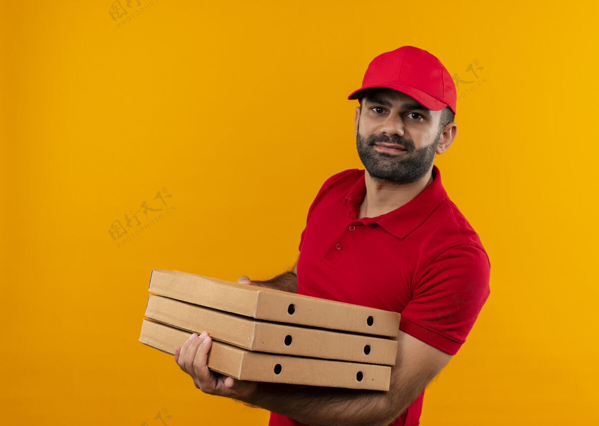 盒子留着胡须的送货员穿着红色制服 戴着帽子 手里拿着一叠披萨盒 站在橙色的墙上 看上去很自信站着叠着拿着