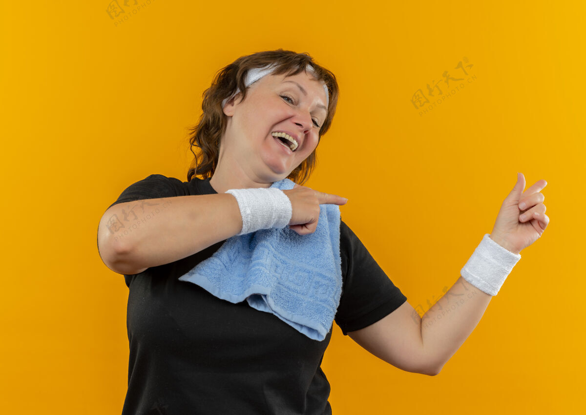 欢呼一位中年运动女性 身穿黑色t恤 头戴头巾 肩上挎着毛巾 面带微笑 手指指向站在橙色墙上的一边年龄中年手指