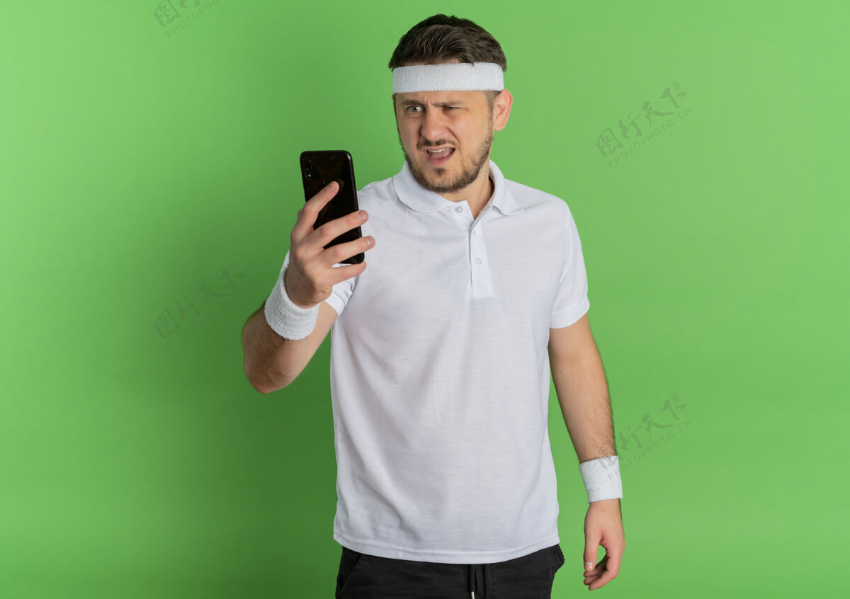 人一个身穿白衬衫 戴着头巾的年轻人站在绿色的墙上 神情困惑地看着手机屏幕表情手机头带