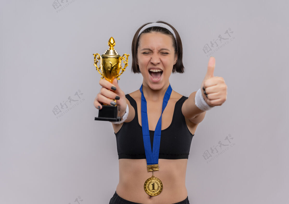 周围一位身穿运动服 脖子上挂着金牌 手持奖杯的年轻健身女士 为自己的成功而高兴和兴奋 她站在白墙上竖起大拇指健身展示女性
